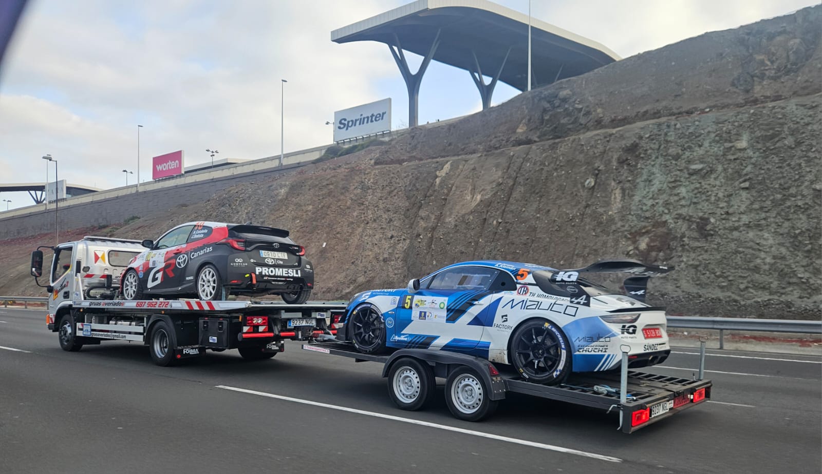 Grúa transportando un coche de rally y remolcando otro coche de rally azul por la autovía
