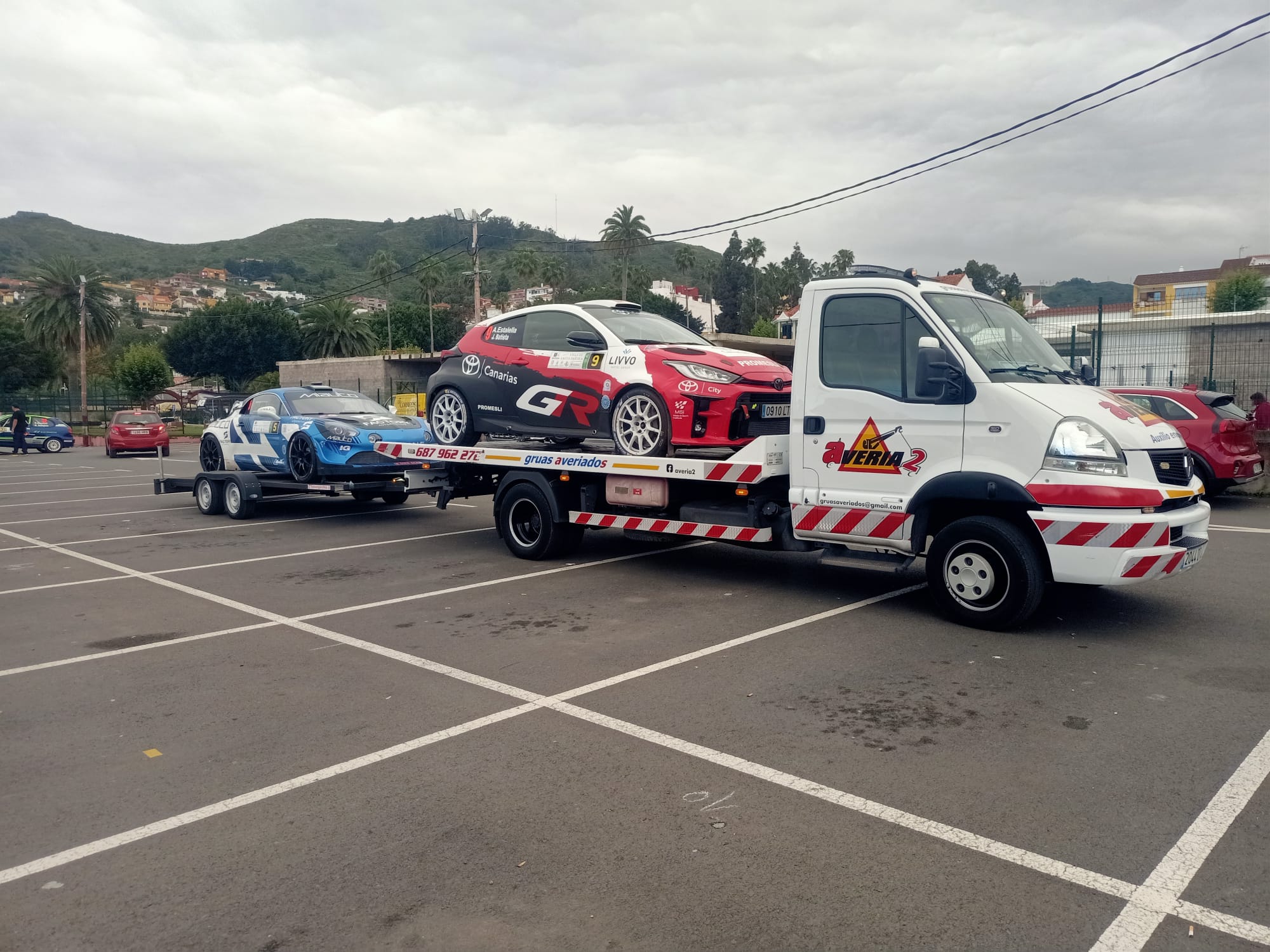 Grúa transportando un coche de rally y remolcando otro coche de rally con colinas de fondo