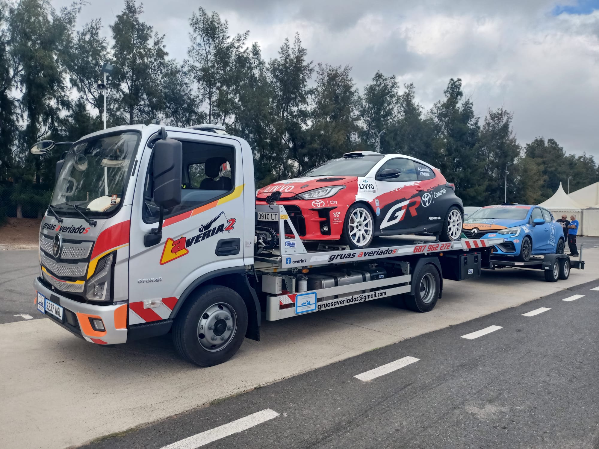 Grúa transportando un coche de rally Toyota GR y remolcando un Renault Clio de rally azul